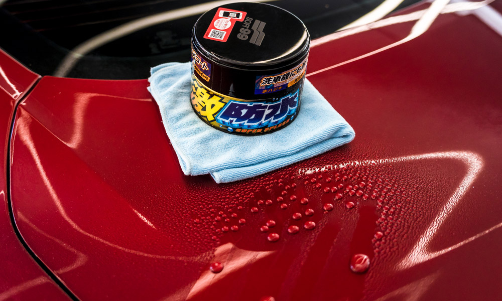 Photo of Soft99 car wax Water Block Super Splash being placed on a microfibre on the rear panel of a red sport car. Next to the wax, round water droplets are visible, signifying the wax's strong hydrophobic properties.